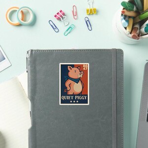 May include: A sticker featuring a cartoon pig wearing a blue bandana and a bandage over its mouth. The sticker has the text "QUIET PIGGY" and is set against a navy blue and orange background. The sticker is on a gray notebook.