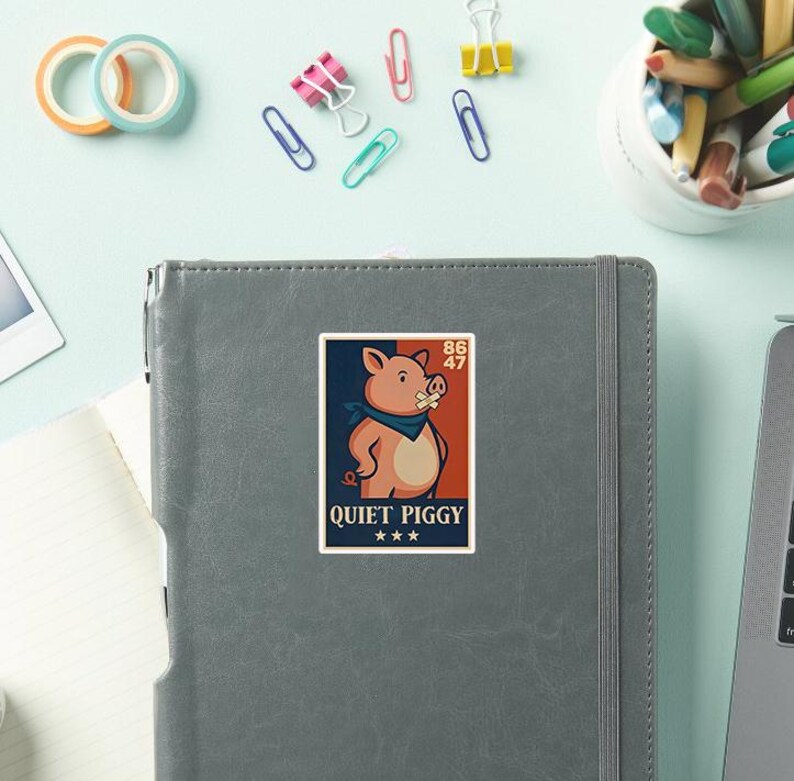 May include: A sticker featuring a cartoon pig wearing a blue bandana and a bandage over its mouth. The sticker has the text "QUIET PIGGY" and is set against a navy blue and orange background. The sticker is on a gray notebook.
