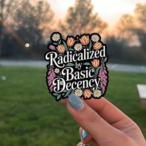 May include: A black sticker with the text "Radicalized by Basic Decency" in white script. The sticker is decorated with orange tulips, white daisies, and pink flowers. The sticker is held up by a hand with blue nail polish.