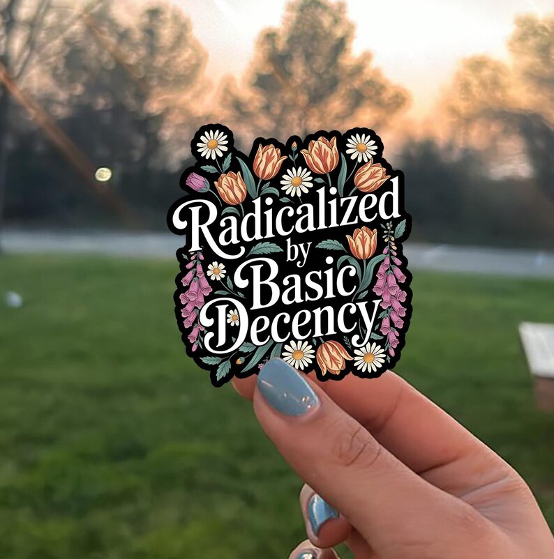 May include: A black sticker with the text "Radicalized by Basic Decency" in white script. The sticker is decorated with orange tulips, white daisies, and pink flowers. The sticker is held up by a hand with blue nail polish.