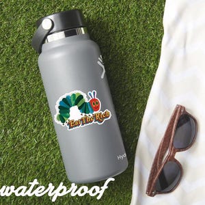 May include: A gray water bottle with a black lid and a colorful sticker featuring a cartoon caterpillar with the text "Eat the Rich." Wooden-framed sunglasses rest on a white cloth, all on a green grassy surface. The word "waterproof" is at the bottom.