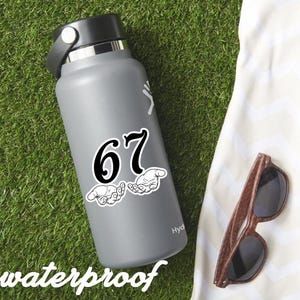 May include: A gray water bottle with a black lid and a white sticker featuring the number 67 and two hands. The word "waterproof" is at the bottom. Brown sunglasses rest on a white towel on a green surface.
