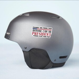 May include: A gray ski helmet with a black chin strap. A sticker on the helmet reads "CLEARLY WE TOOK THIS ANYONE CAN BE PRESIDENT THING A BIT TOO FAR" in red, white, and blue.
