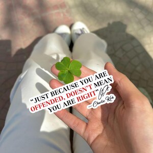 May include: A hand holding a four-leaf clover with a sticker that reads "JUST BECAUSE YOU ARE OFFENDED, DOESN'T MEAN YOU ARE RIGHT" by Charlie Kirk. The clover is bright green.