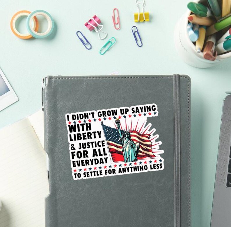 Peut inclure: Un autocollant avec le texte "I DIDN'T GROW UP SAYING WITH LIBERTY & JUSTICE FOR ALL EVERYDAY TO SETTLE FOR ANYTHING LESS" repr&eacute;sentant la Statue de la Libert&eacute; et un drapeau am&eacute;ricain.