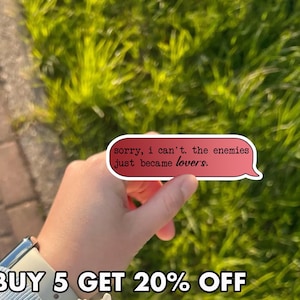 May include: A red speech bubble sticker with the text "sorry, i can't. the enemies just became lovers." is held up against a background of green grass. The sticker has a white border and a light pink interior.