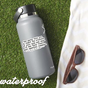 May include: A gray insulated water bottle with a black lid and a white sticker featuring a quote from George Orwell's "1984." The sticker reads, "the party told you to reject the evidence of your eyes and ears..." Wooden-framed sunglasses rest on a white cloth.