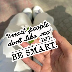 May include: A white sticker with the text "smart people don't like me" and "BE SMART." The sticker is held in a hand, with white pants and shoes visible in the background.