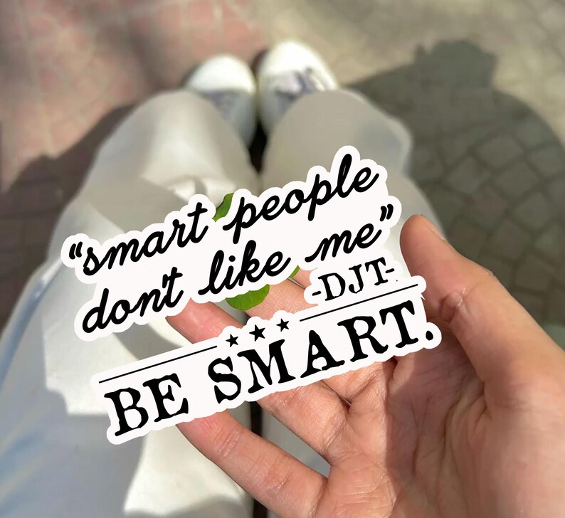 May include: A white sticker with the text "smart people don't like me" and "BE SMART." The sticker is held in a hand, with white pants and shoes visible in the background.