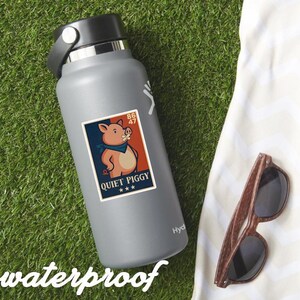 May include: A gray water bottle with a "Quiet Piggy" sticker featuring a cartoon pig wearing a bandana. The sticker has a red, white, and blue color scheme. Wooden-framed sunglasses rest on a white towel, all on a green grassy surface.