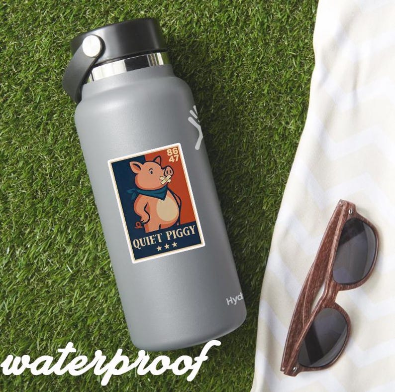 May include: A gray water bottle with a "Quiet Piggy" sticker featuring a cartoon pig wearing a bandana. The sticker has a red, white, and blue color scheme. Wooden-framed sunglasses rest on a white towel, all on a green grassy surface.