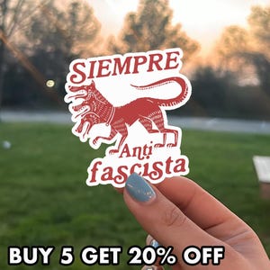 May include: A white sticker with a red illustration of a three-headed dog and the words "SIEMPRE Anti fascista". The sticker is held in a hand with a blurred background of green grass and trees.