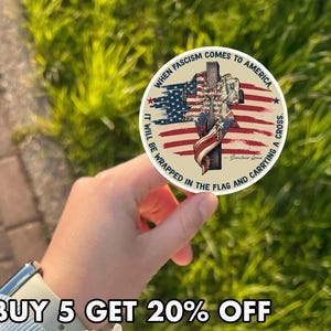 May include: A round sticker with an American flag design and the text "WHEN FASCISM COMES TO AMERICA IT WILL BE WRAPPED IN THE FLAG AND CARRYING A CROSS." The sticker has a cross and a soldier's silhouette.