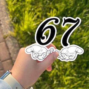 May include: A sticker featuring the numbers 67 in black with a white outline, held by a hand. The numbers are positioned above two stylized hands. The background is blurred green grass.