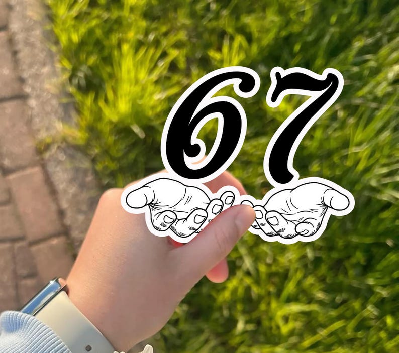 May include: A sticker featuring the numbers 67 in black with a white outline, held by a hand. The numbers are positioned above two stylized hands. The background is blurred green grass.