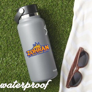 May include: A gray insulated water bottle with a black cap and a strap. The bottle has a sticker with the text "ZOHRAN for NEW YORK CITY" in blue, orange, and yellow. Wooden-framed sunglasses rest on a white towel on a green surface. The word "waterproof" is at the bottom.