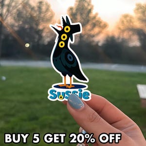 May include: A black sticker featuring a stylized bird-like creature with yellow eyes and a blue circular design. The sticker has the name "Sussie" in blue and white text. The sticker is held up by a hand with blue nail polish.