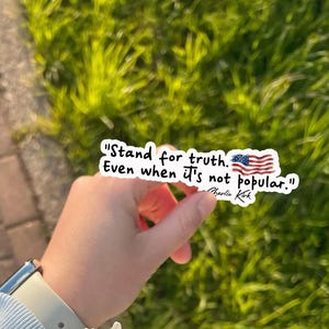 May include: A white sticker with the text "Stand for truth. Even when it's not popular." and an American flag graphic. The sticker is held against a background of green grass and a brick walkway.