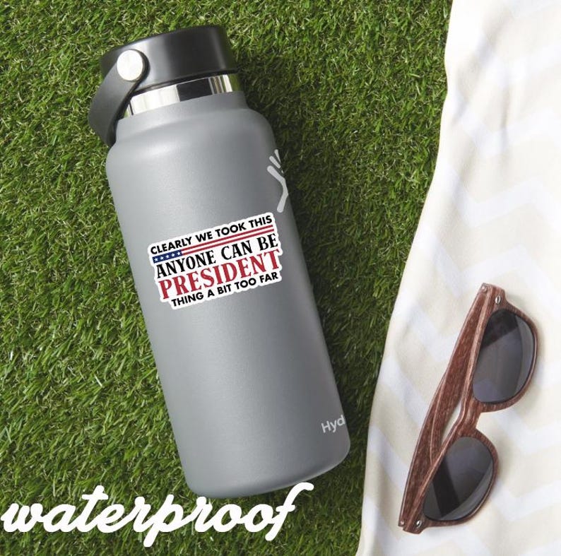 May include: A gray water bottle with a black lid and a sticker that reads "CLEARLY WE TOOK THIS ANYONE CAN BE PRESIDENT THING A BIT TOO FAR." The bottle is next to a pair of wooden-framed sunglasses.