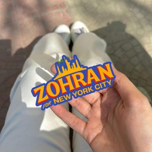 May include: A colorful sticker with the text "ZOHRAN FOR NEW YORK CITY" in bold orange and yellow letters, with a blue skyline graphic. The sticker is held in a person's hand.