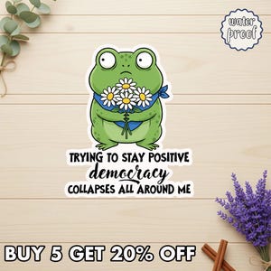 May include: A green frog sticker holding daisies, wearing a blue bandana, with the text "TRYING TO STAY POSITIVE democracy COLLAPSES ALL AROUND ME." The sticker is labeled "water proof."