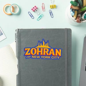 May include: Gray notebook with the text "ZOHRAN FOR NEW YORK CITY" in blue and yellow. The notebook is on a light blue surface with office supplies, including paper clips and tape.