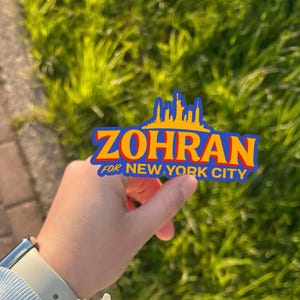 May include: A sticker with the text "ZOHRAN FOR NEW YORK CITY" in blue and orange, with a skyline graphic. The sticker is held up against a background of green grass.