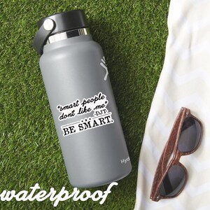 May include: A gray Hydro Flask water bottle with a black lid and handle. A white sticker on the bottle reads "smart people don't like me" and "BE SMART." The word "waterproof" is written at the bottom. Wooden sunglasses rest on a white towel.