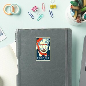 May include: A sticker featuring a stylized portrait with red, white, and blue colors. The word "FASCIST" is printed in a dark blue box below the portrait. The sticker is on a gray notebook.