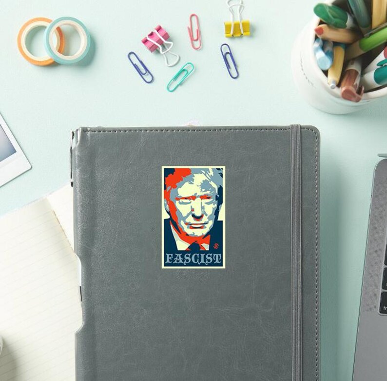 May include: A sticker featuring a stylized portrait with red, white, and blue colors. The word "FASCIST" is printed in a dark blue box below the portrait. The sticker is on a gray notebook.