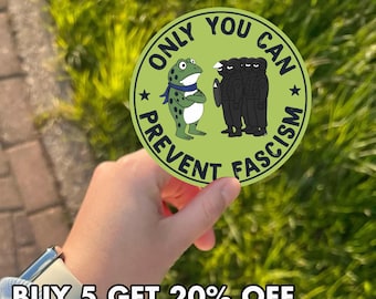 May include: A round, green sticker with the text "ONLY YOU CAN PREVENT FASCISM" encircling a cartoon frog and a group of figures. The frog wears a blue scarf. The sticker is held against a background of green foliage.
