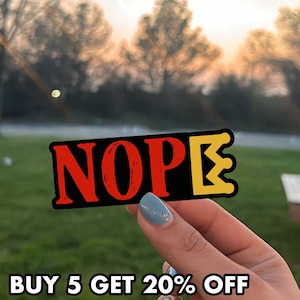 May include: A rectangular sticker with the word "NOPE" in red letters on a black background. The letter "E" is yellow with a crown design. The sticker is held up against a blurred outdoor background.
