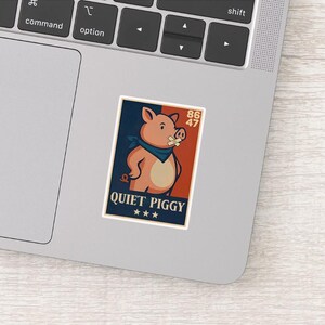 May include: A sticker featuring a cartoon pig with a blue bandana and a bandage over its mouth. The background is split between navy blue and red, with the text "QUIET PIGGY" and three stars at the bottom. Numbers "86" and "47" are in the upper right.