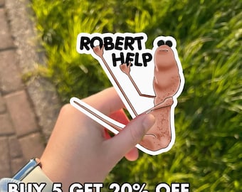 May include: A sticker featuring a cartoon character with the text "ROBERT HELP" in a black and white font. The character is a brown, elongated shape with arms and sunglasses. The sticker is held against a background of green grass.