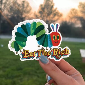 May include: A colorful sticker featuring a cartoon caterpillar with a red face and blue and green body segments. The sticker has the text "Eat The Rich" in a playful, multi-colored font.