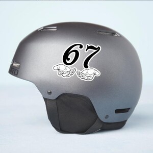 May include: A gray ski helmet with a black chin strap. A sticker with the number "67" in black, with two hands cupped underneath, is affixed to the side of the helmet. The helmet has the brand name "GIRO" on the side.