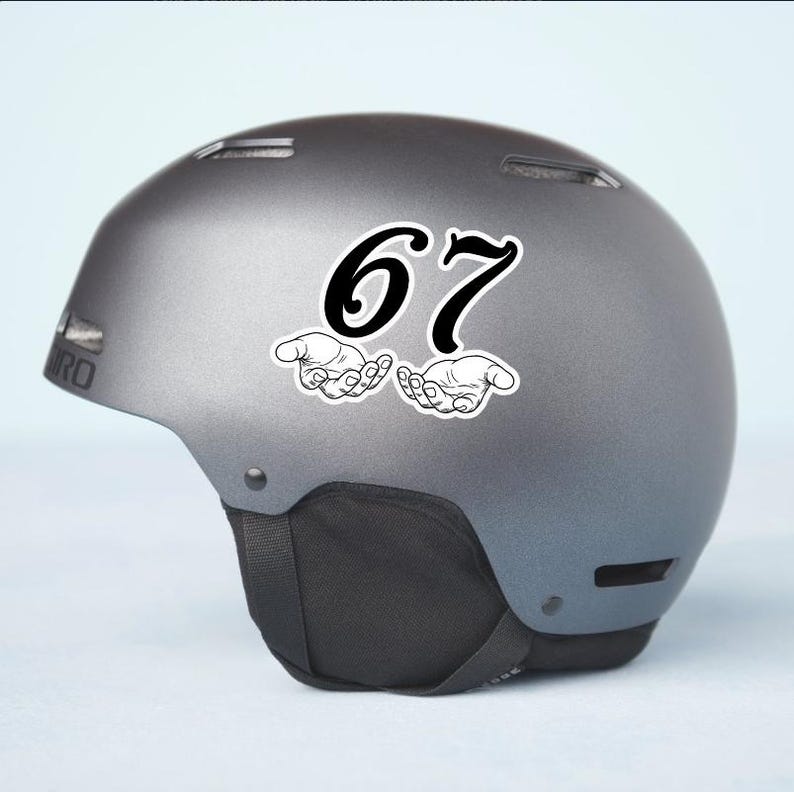 May include: A gray ski helmet with a black chin strap. A sticker with the number "67" in black, with two hands cupped underneath, is affixed to the side of the helmet. The helmet has the brand name "GIRO" on the side.