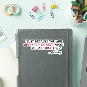 May include: A gray notebook with the quote "JUST BECAUSE YOU ARE OFFENDED, DOESN'T MEAN YOU ARE RIGHT" by Charlie Kirk. The image also shows colorful office supplies like paper clips and tape.