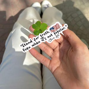 May include: A white sticker with the quote "Stand for truth. Even when it's not popular." and an American flag graphic. A green four-leaf clover is also visible. The sticker is held in a hand.