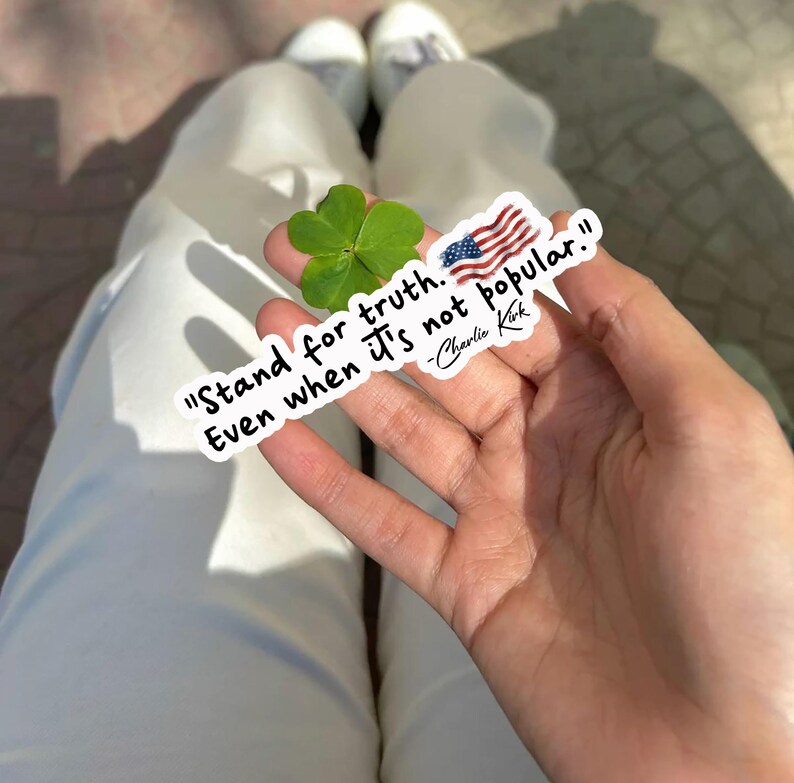 May include: A white sticker with the quote "Stand for truth. Even when it's not popular." and an American flag graphic. A green four-leaf clover is also visible. The sticker is held in a hand.