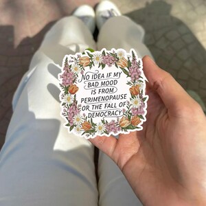May include: A white sticker with a floral border featuring the text: "No idea if my bad mood is from perimenopause or the fall of democracy." The sticker is held in a hand, with white pants and shoes in the background.