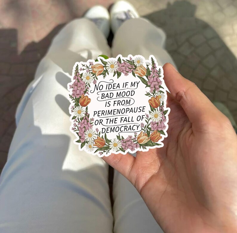 May include: A white sticker with a floral border featuring the text: "No idea if my bad mood is from perimenopause or the fall of democracy." The sticker is held in a hand, with white pants and shoes in the background.