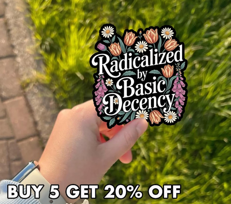 May include: A sticker with the text "Radicalized by Basic Decency" in a floral design with daisies, tulips, and other flowers. The sticker is held in a hand against a background of green grass.