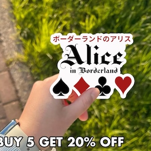 May include: A white sticker with the text "Alice in Borderland" in black gothic font, with Japanese text above. The sticker includes playing card suit symbols: spades, diamonds, clubs, and hearts. The background is blurred green foliage.