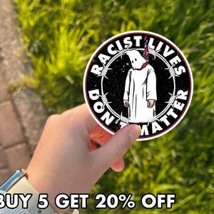May include: A round sticker with a black background and white text that reads "RACIST LIVES DON'T MATTER". The sticker features a white-robed figure with a pointed hood inside a barbed wire circle.
