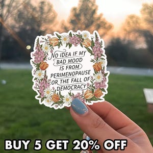 May include: A white sticker with a floral border featuring the text: "NO IDEA IF MY BAD MOOD IS FROM PERIMENOPAUSE OR THE FALL OF DEMOCRACY." The sticker is decorated with orange tulips, white daisies, and purple flowers.