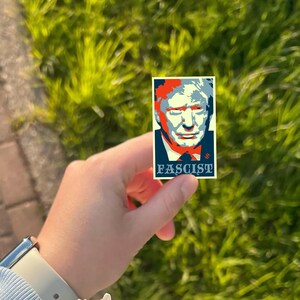 May include: A rectangular sticker featuring a stylized portrait with red, blue, and white colors. The word "FASCIST" is printed in white at the bottom. The sticker is held against a background of green grass.