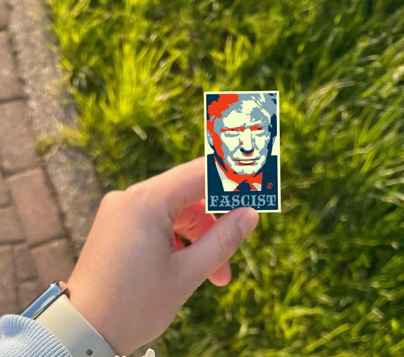 May include: A rectangular sticker featuring a stylized portrait with red, blue, and white colors. The word "FASCIST" is printed in white at the bottom. The sticker is held against a background of green grass.
