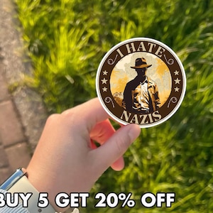 May include: A round sticker with the text "I HATE NAZIS" and an illustration of a person in a hat and jacket. The sticker is brown and tan with a star design around the edge. The text "BUY 5 GET 20% OFF" is at the bottom.