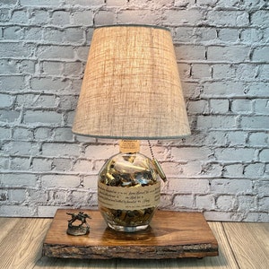 May include: A unique table lamp with a beige fabric shade and a clear glass base filled with brass bullet casings. The lamp sits on a live edge wooden base, with a small metal horse figurine. The background is a white brick wall.